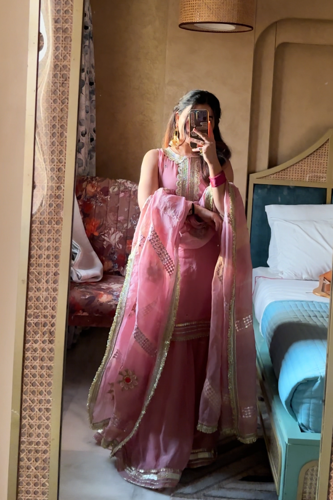 Person wearing a pink traditional outfit in a room with a bed and lamp.