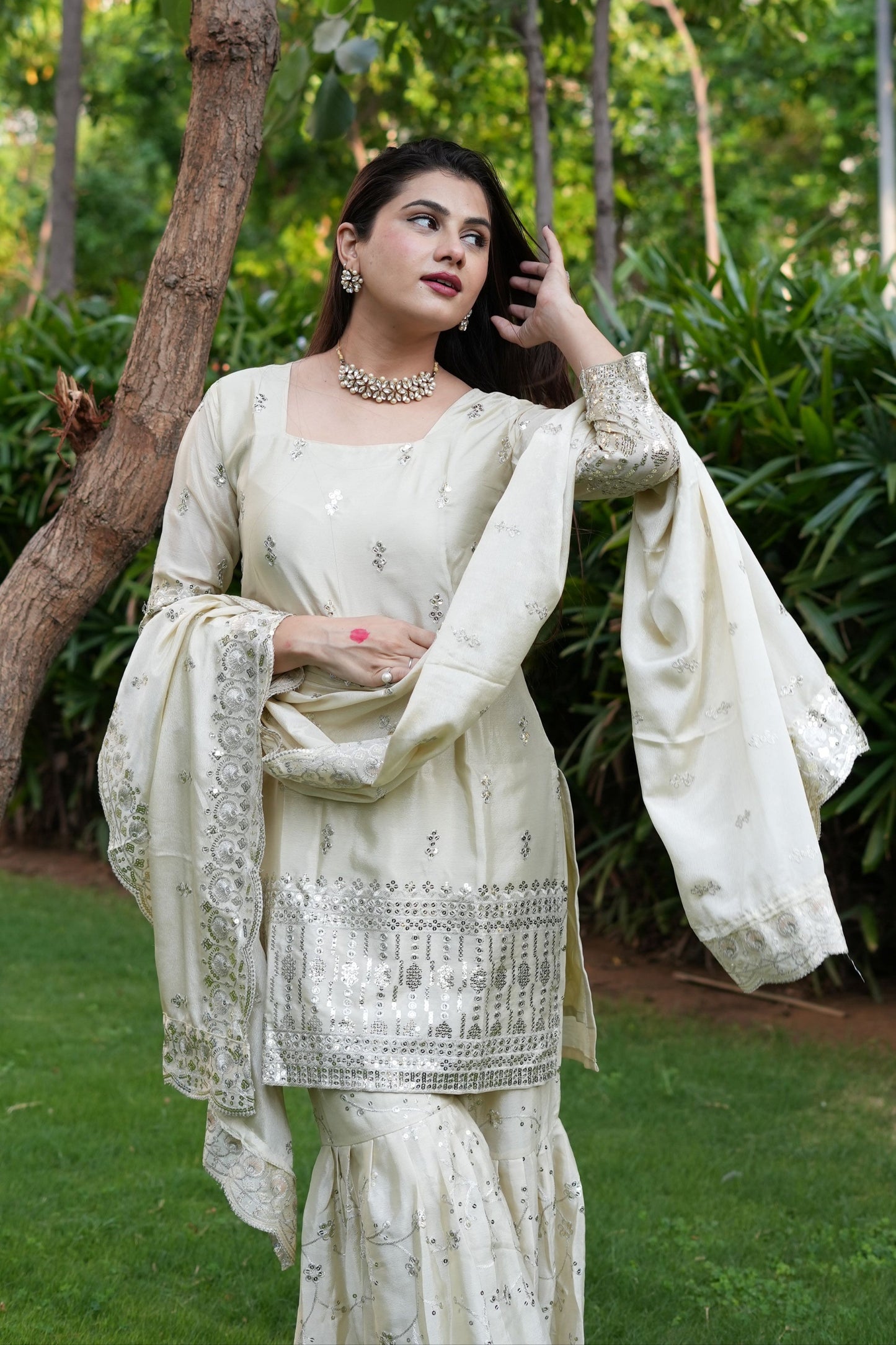Woman in a white traditional outfit with embroidery, standing outdoors with greenery.