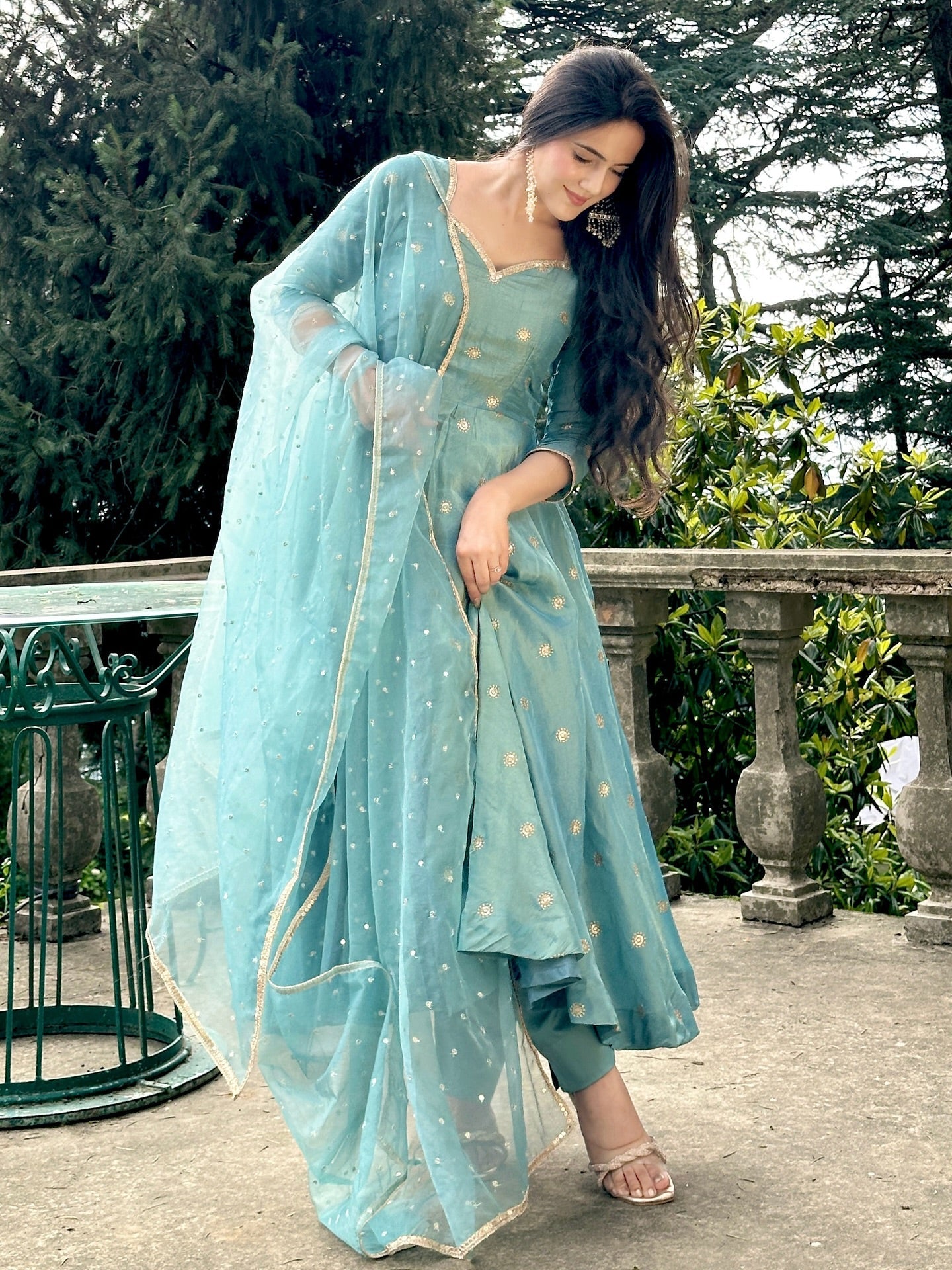 Woman in a light blue traditional outfit standing outdoors with greenery in the background