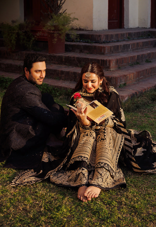Man and woman sitting on grass, man holding a book and flower, woman in traditional attire.