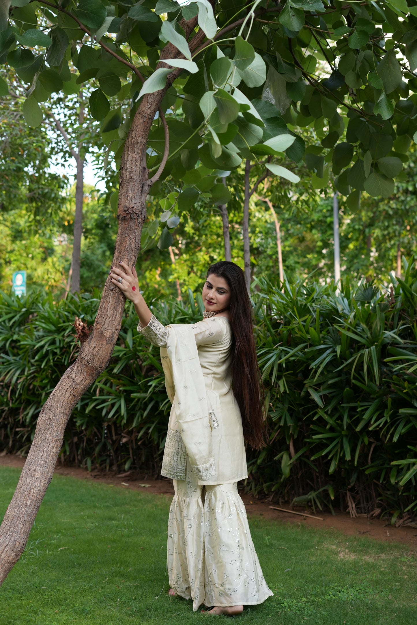 Woman in a white traditional outfit standing under a tree with greenery around