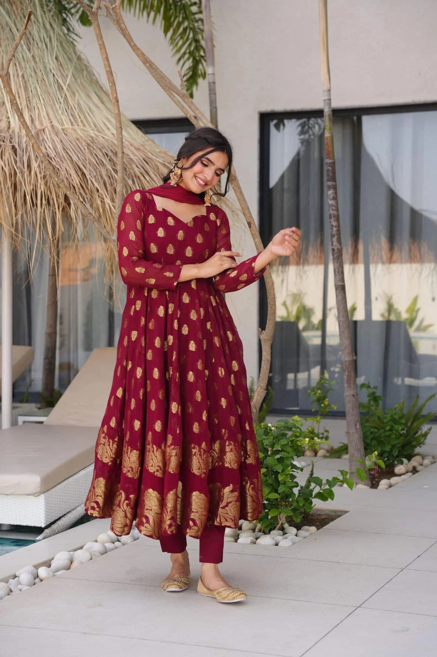 Woman in a red and gold dress standing in an outdoor setting with plants and a building in the background.