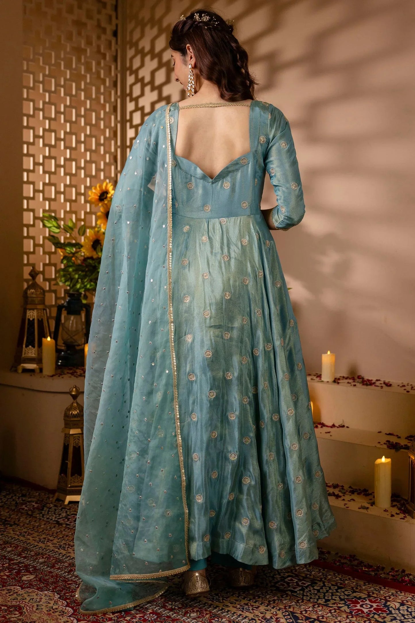 Woman in a light blue traditional outfit with a floral pattern, standing in a decorated room.