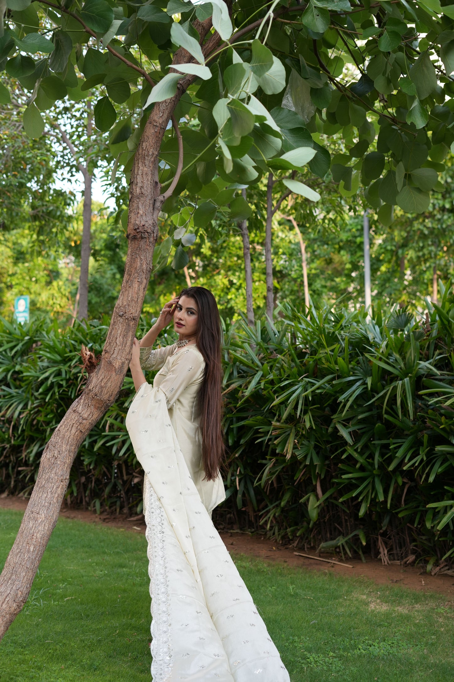 Woman in a white dress standing next to a tree in a green outdoor setting