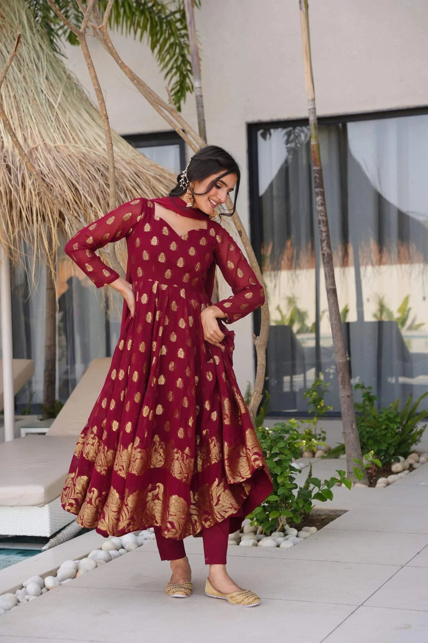 Woman in a maroon and gold traditional outfit standing outdoors with plants and a building in the background.