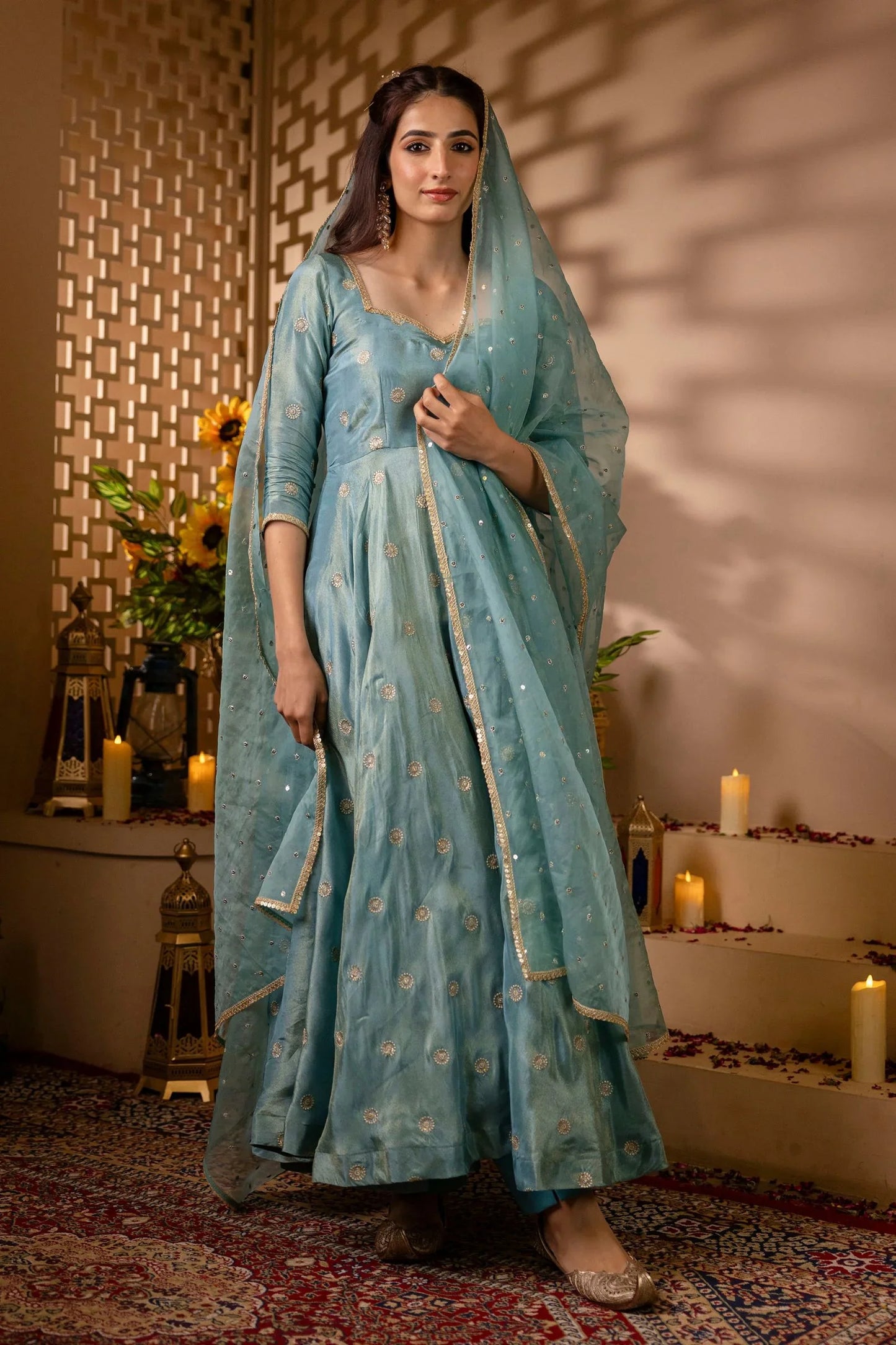 Woman in a light blue traditional outfit standing in a decorated room with candles and flowers.