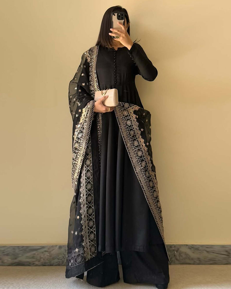 Person wearing a black outfit with an embroidered abaya against a beige wall.