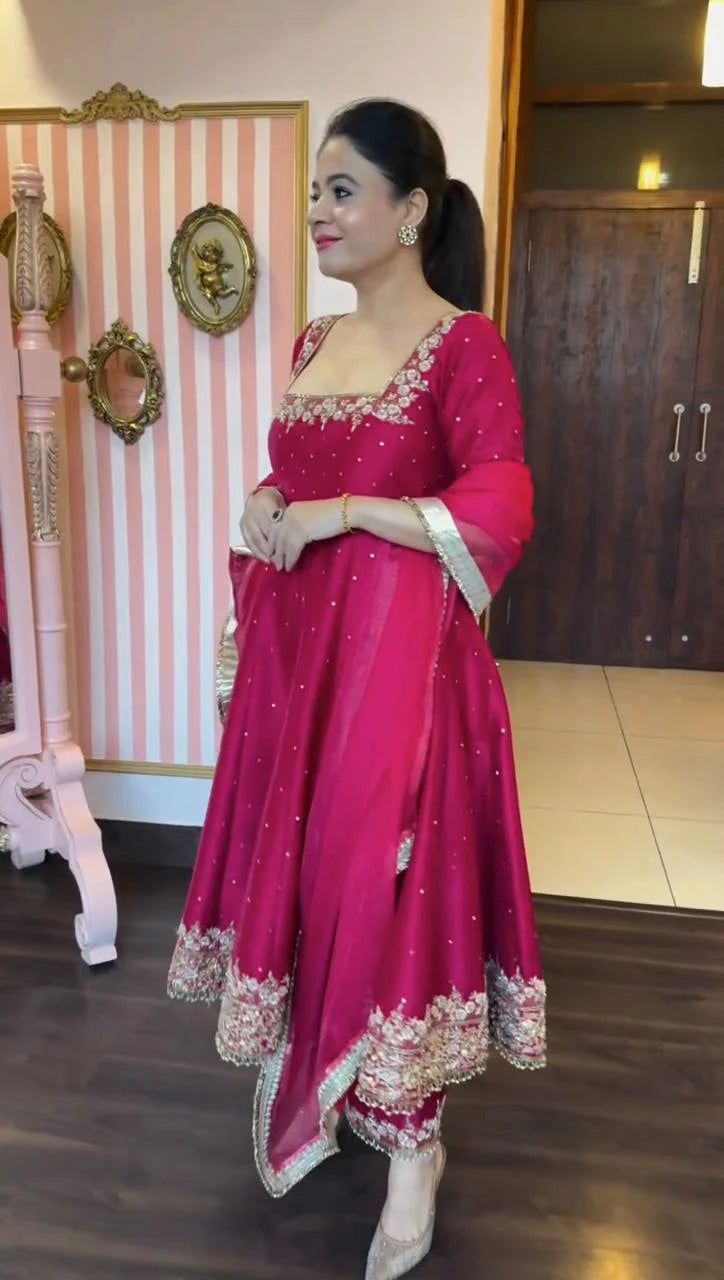 Woman in a pink traditional outfit standing indoors with decorative elements in the background.