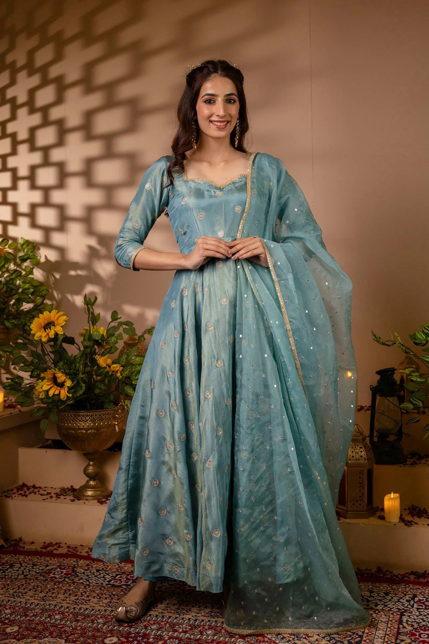 Woman in a light blue traditional outfit standing in a decorated indoor setting with plants and candles.