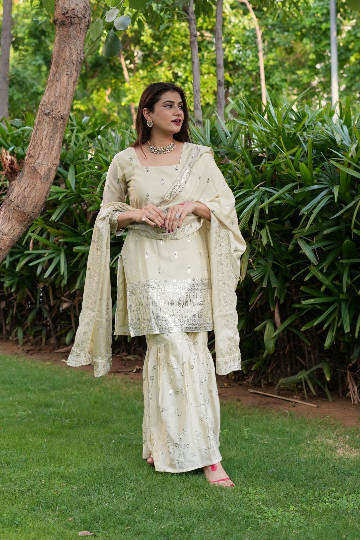 Woman in a light-colored traditional outfit standing in a natural setting with greenery.