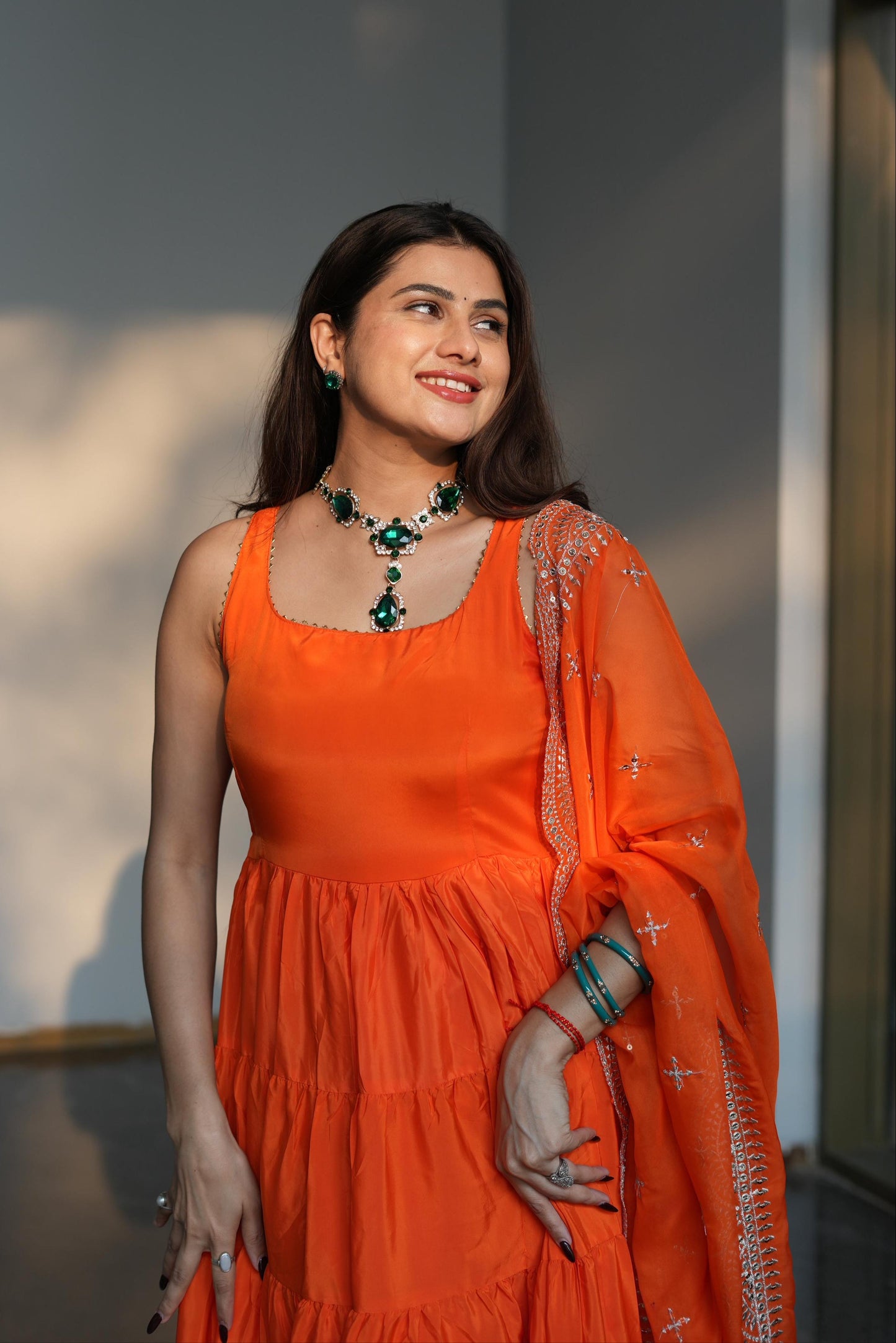 Woman in an orange dress with a green necklace against a neutral background