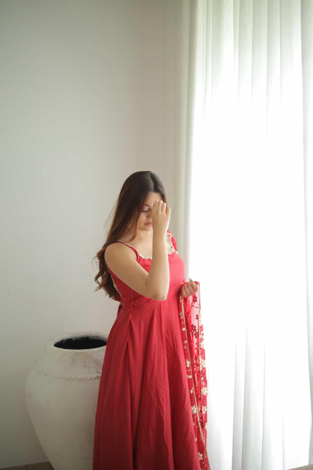 Woman in a red dress standing by a window with white curtains.