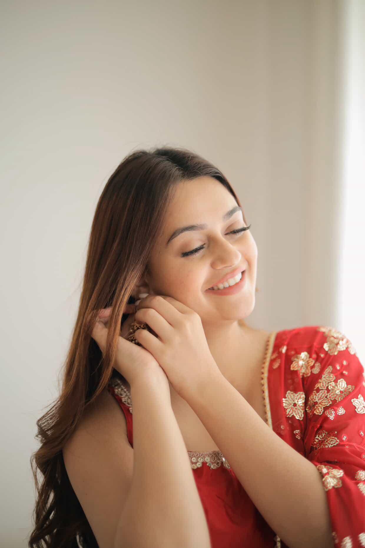 Woman in a red traditional outfit adjusting her earring against a neutral background