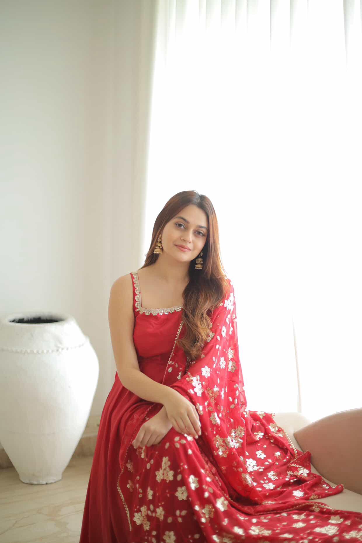 Woman in a red floral dress sitting indoors with a white curtain background