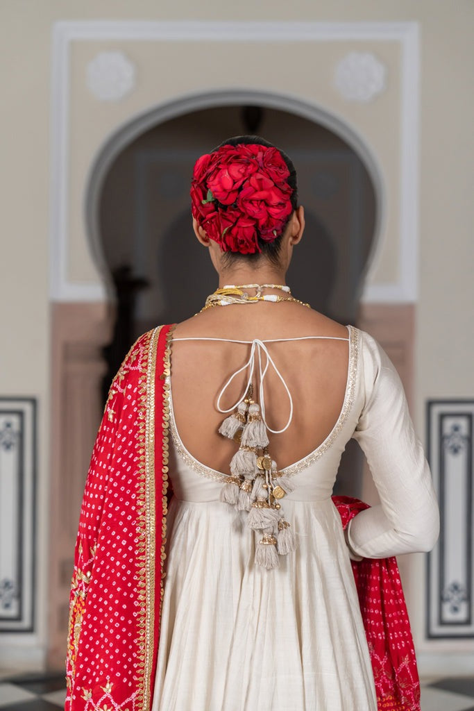 Woman in a white dress with red shawl and floral headpiece, standing in front of an archway.