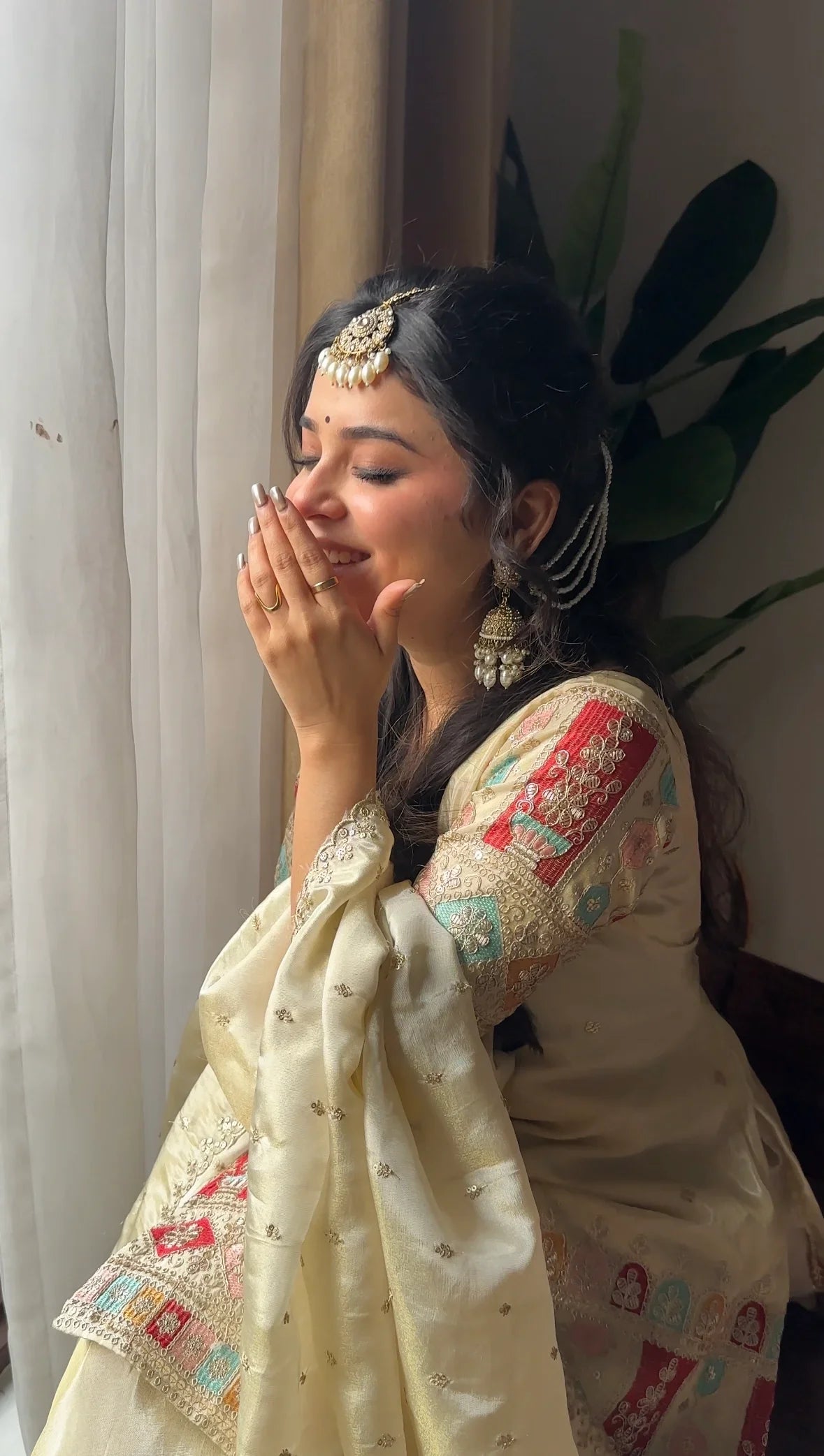 Woman in traditional attire with jewelry, standing indoors near a curtain.