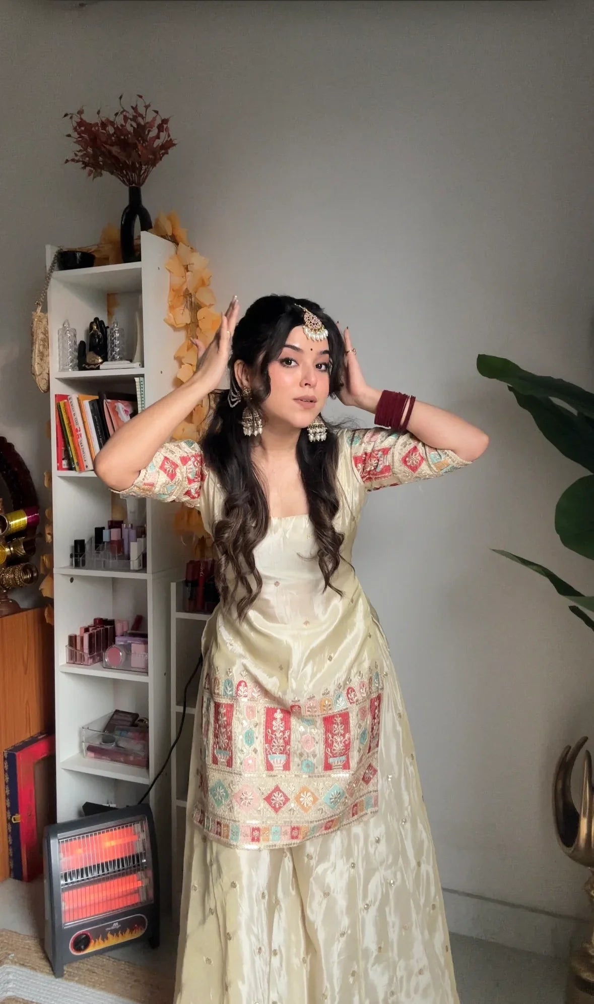Woman in a traditional outfit standing in a room with a bookshelf and decorative items.