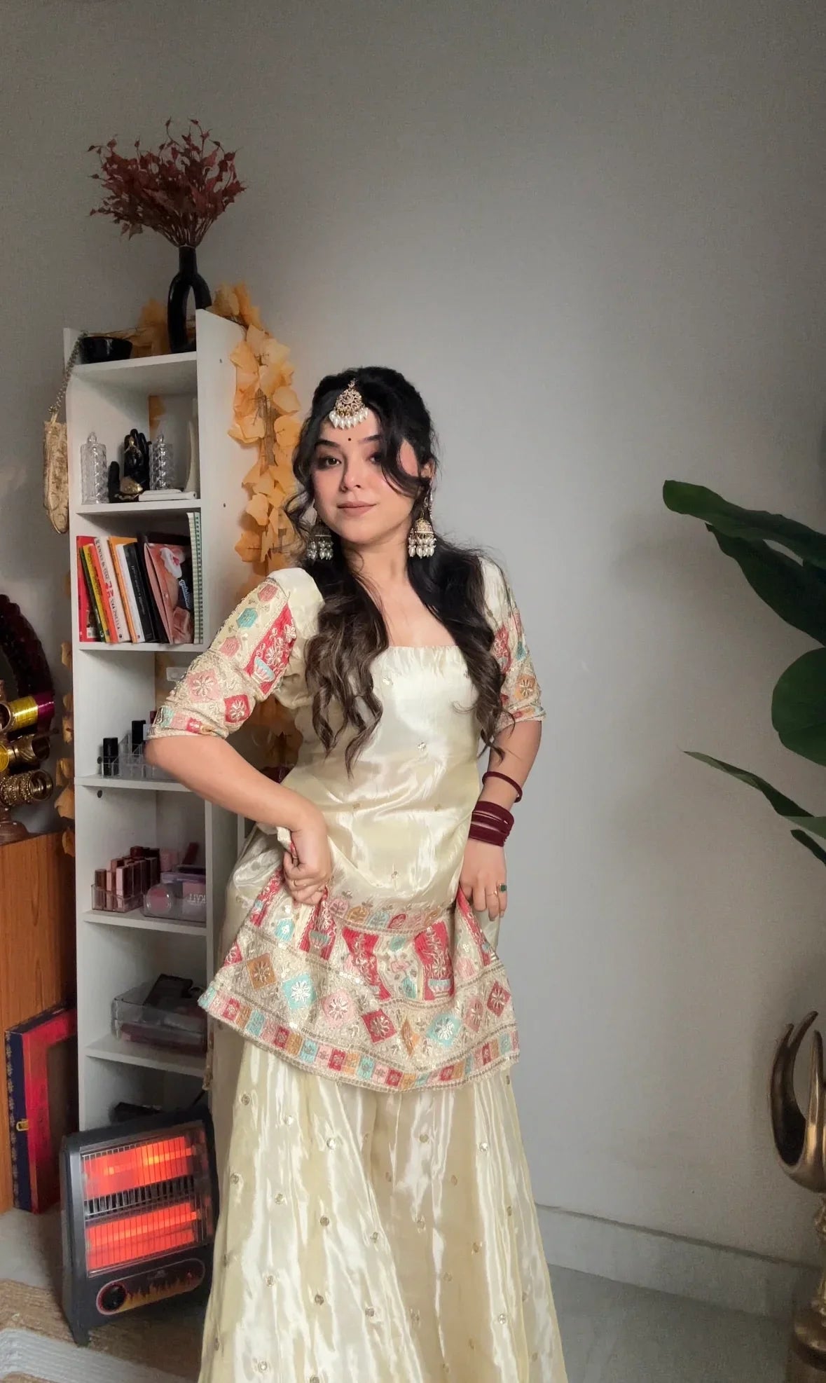 Woman in a traditional outfit standing in a room with a bookshelf and plant.
