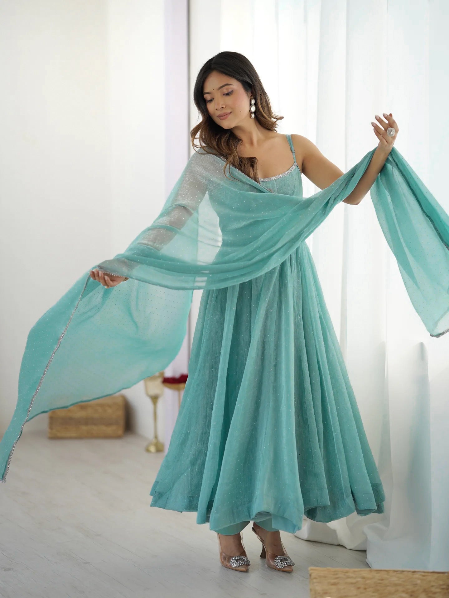 Woman in a long turquoise dress holding a matching scarf against a white curtain background