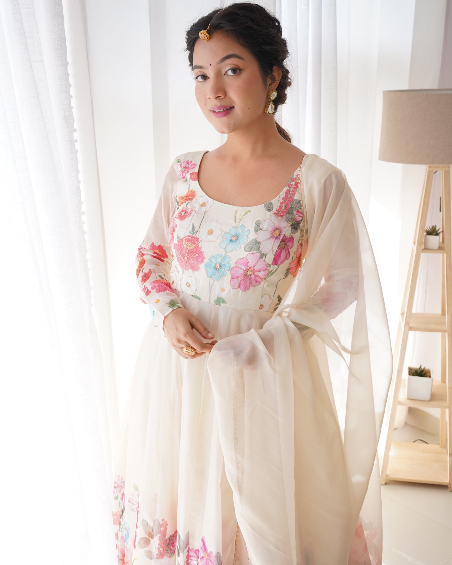 Woman wearing a floral dress with a sheer white overlayer in a bright room.