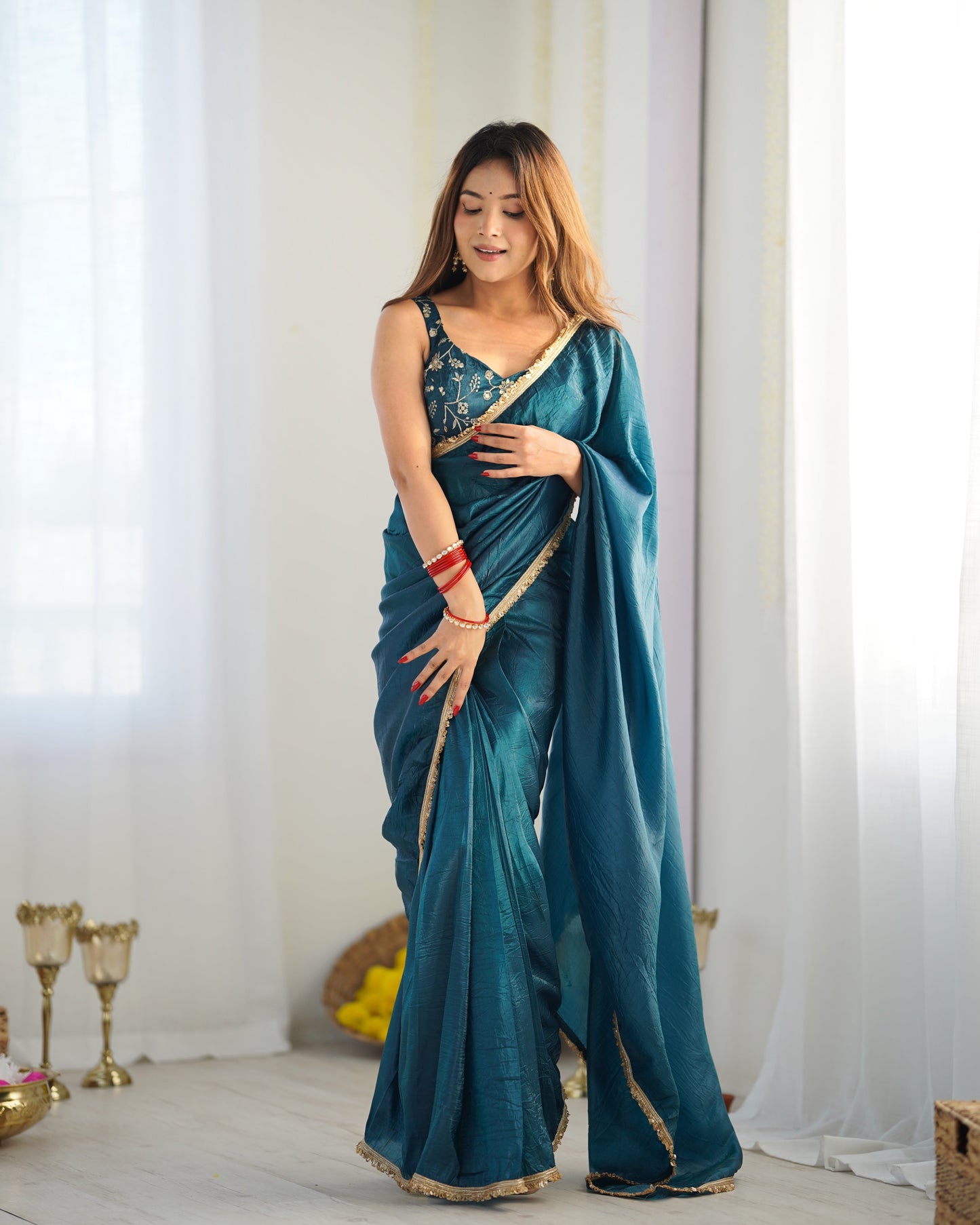 Woman in a teal saree with gold border standing in a well-lit room.