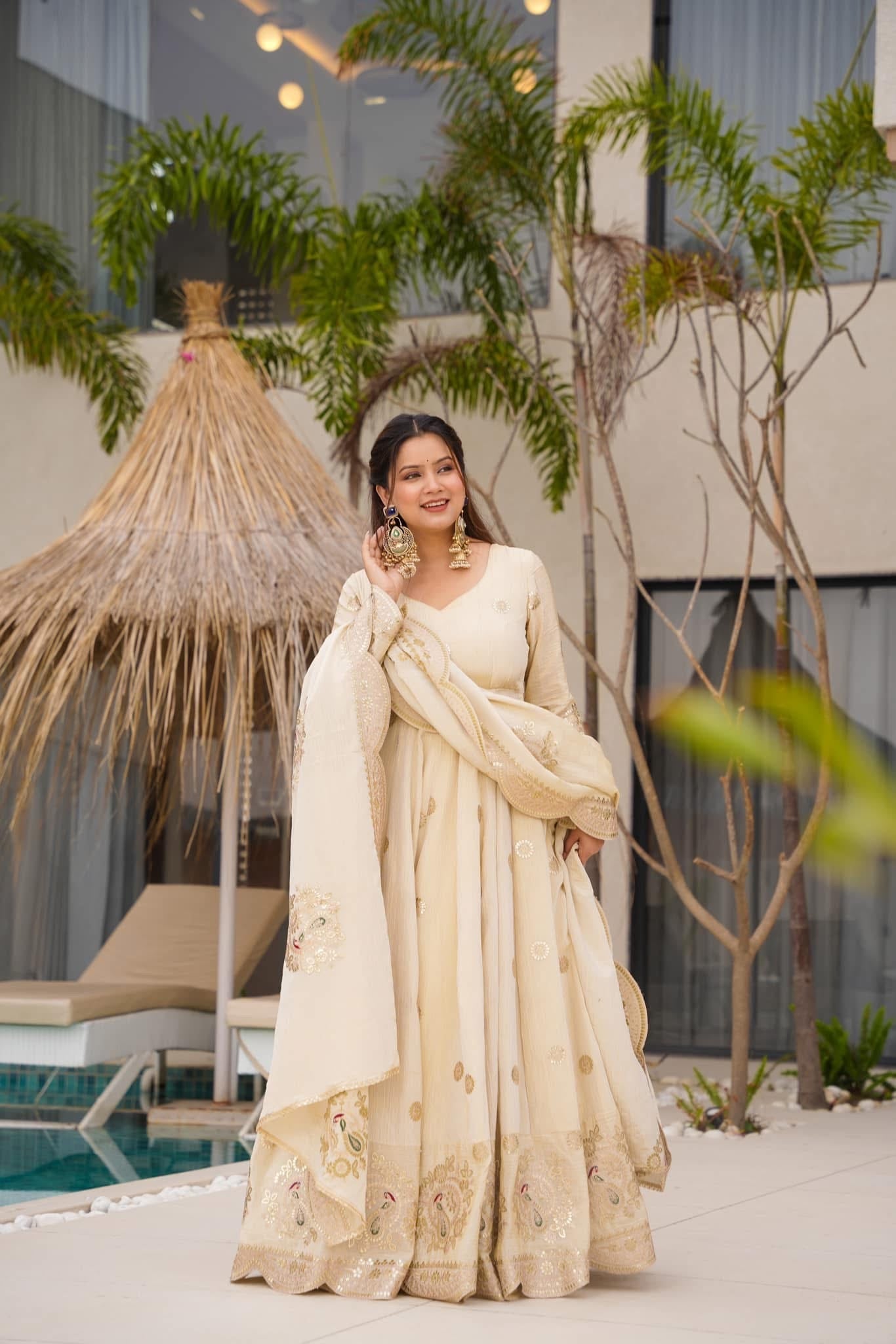 Woman in a cream saree standing by a poolside with plants and a thatched roof structure in the background.