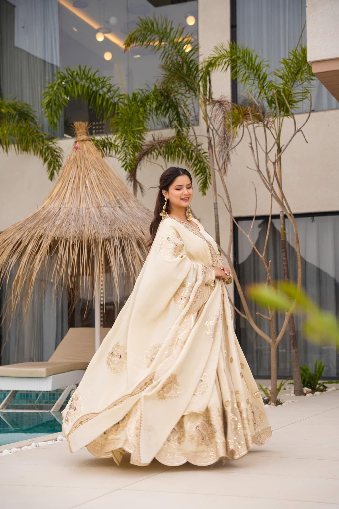 Woman in a traditional outfit standing in an outdoor setting with plants and a pool.
