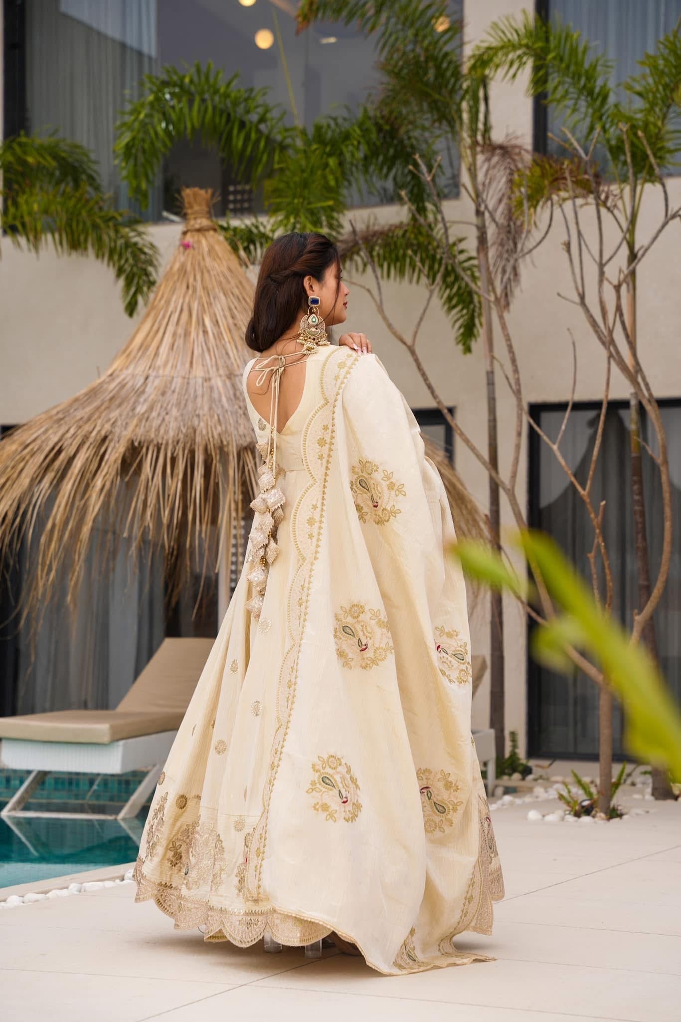 Woman in a cream saree with floral patterns standing outdoors near plants and a building.
