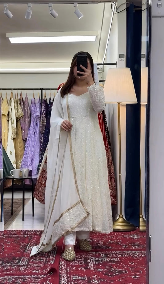 Person wearing a white traditional outfit with gold details in a room with a mirror and clothing rack.