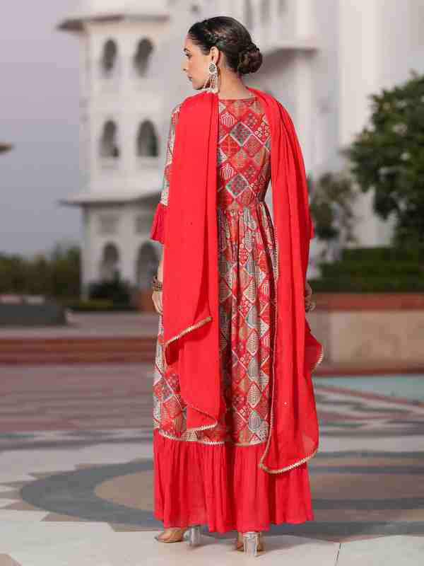Woman in a red traditional outfit with a patterned dress and dupatta, standing in front of a white building.