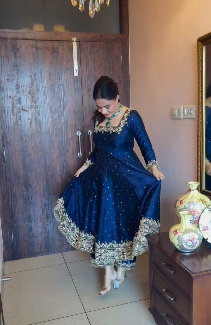 Woman in a blue traditional dress standing in a room with wooden furniture and a decorative vase.