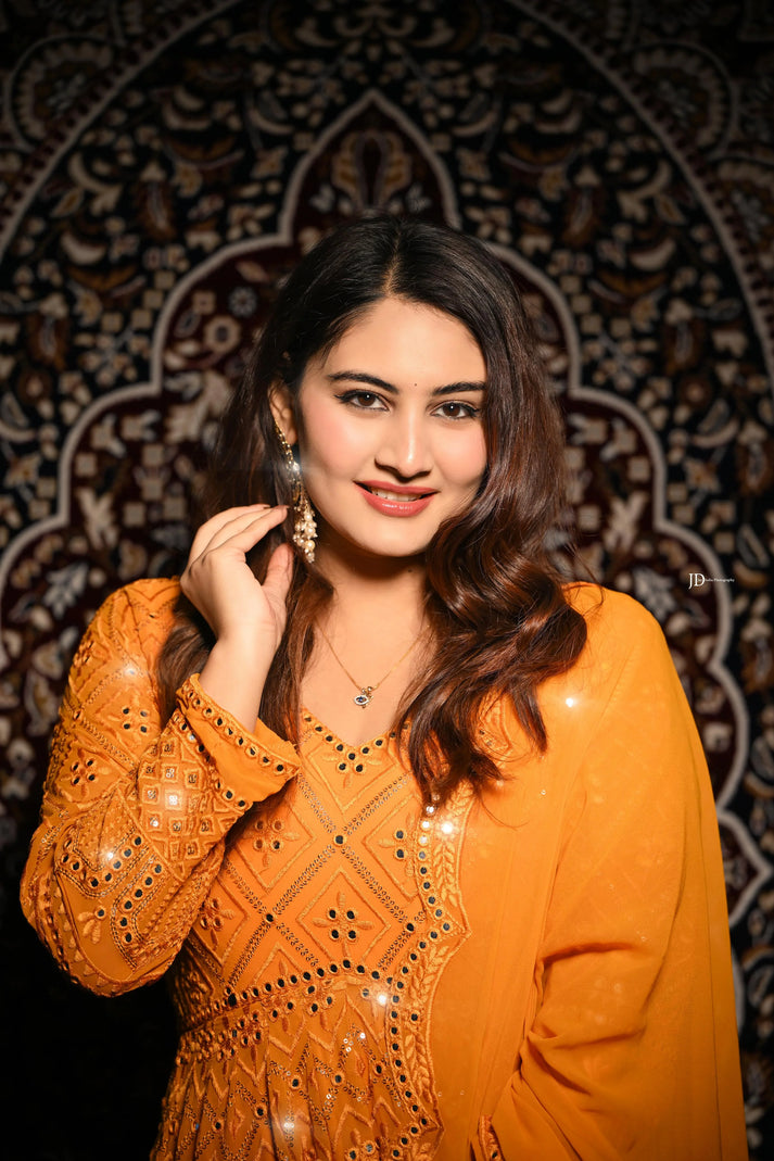 Woman in an orange traditional outfit with intricate patterns against a decorative dark background