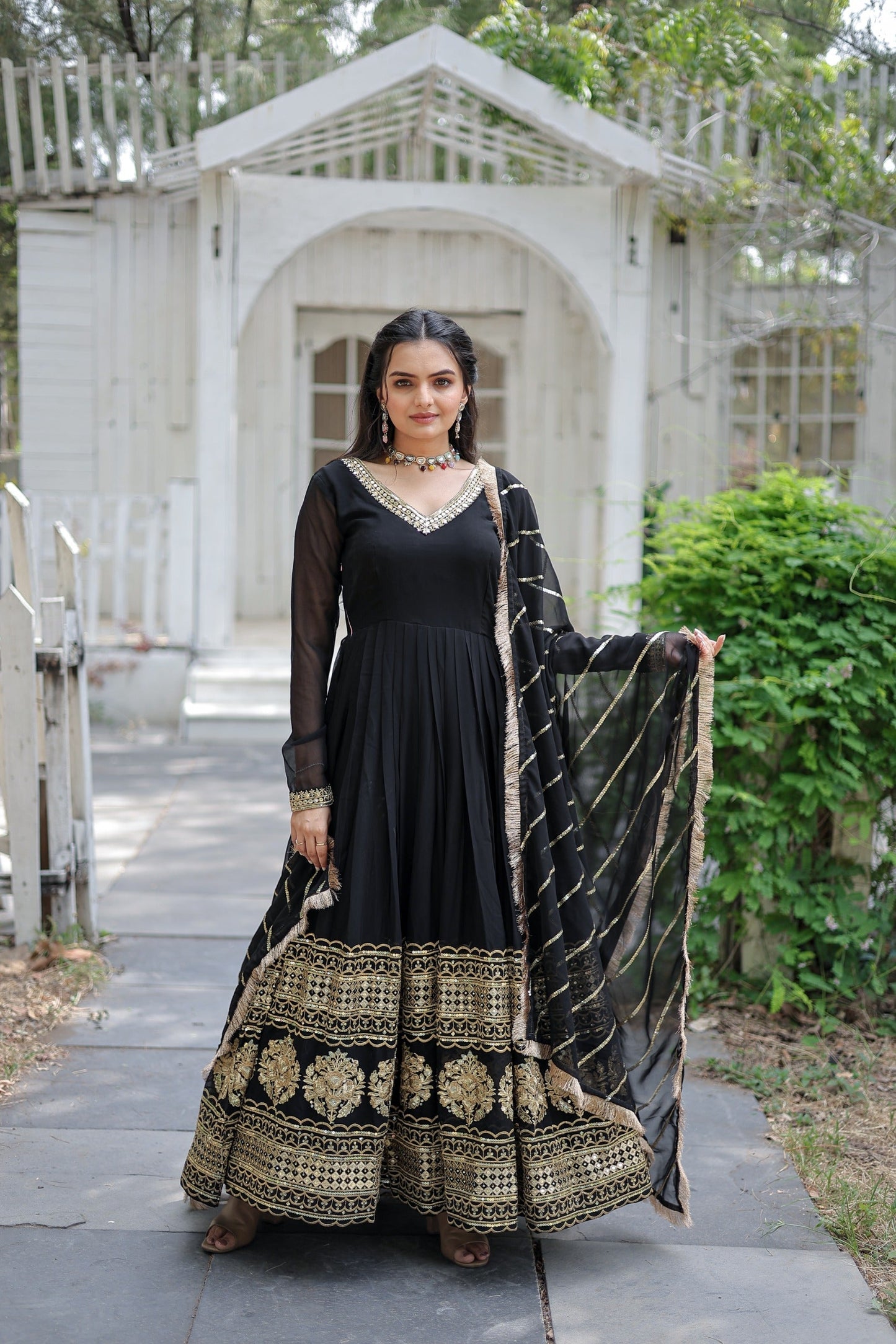 Woman in a black and gold traditional outfit standing outdoors with a white structure and greenery in the background.