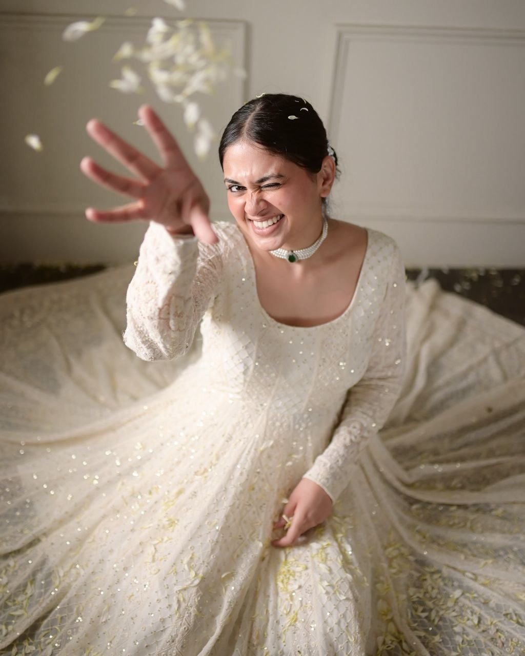 Woman in a white dress with long sleeves, sitting on a white surface with flowers around her.