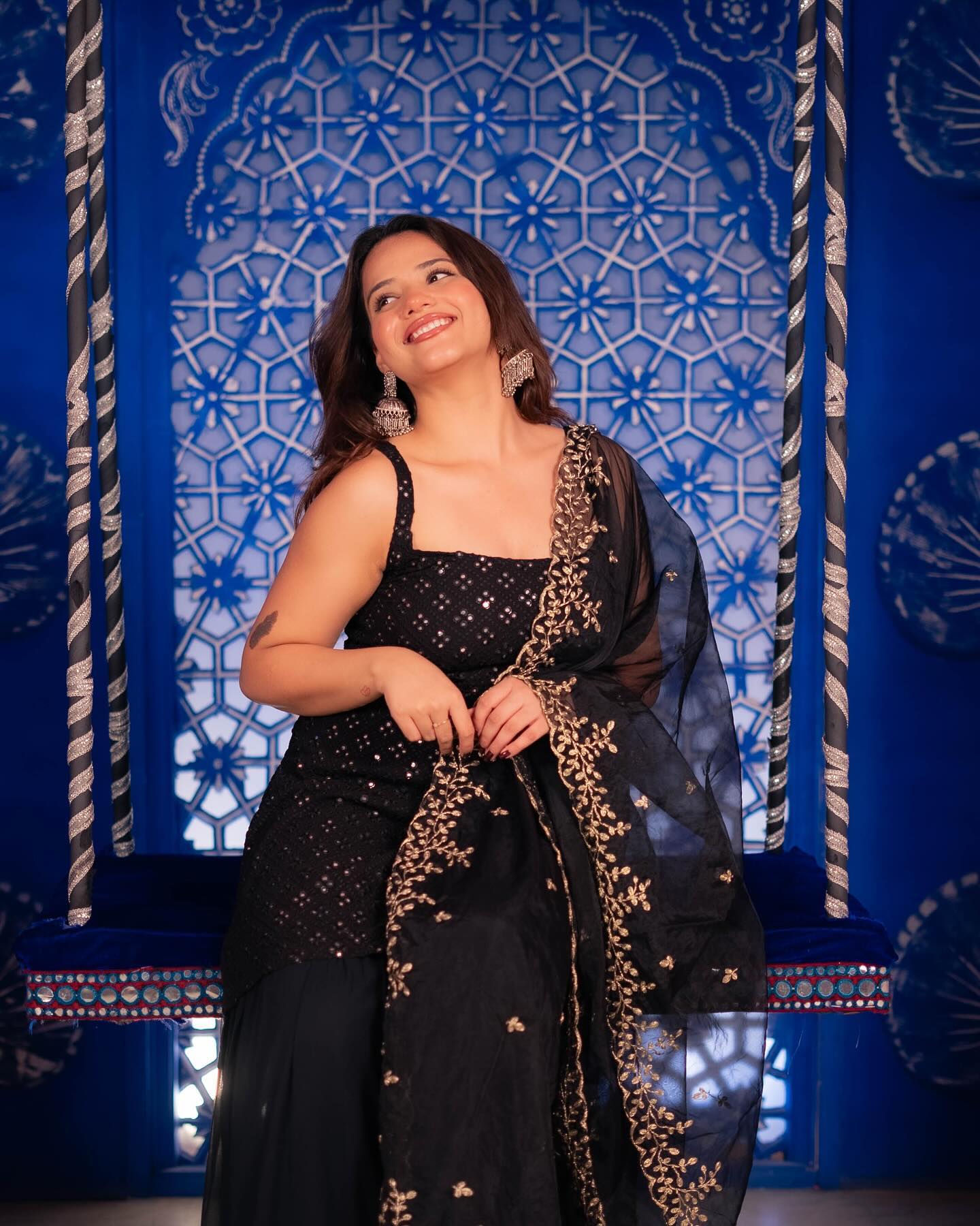Woman in a black traditional outfit with gold details against a blue decorative wall.