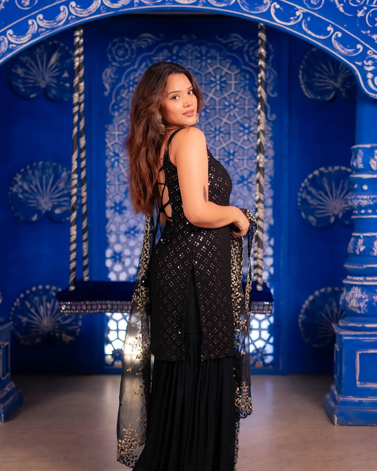 Woman in a black dress with a long, beaded scarf standing in front of a blue wall with white patterns.