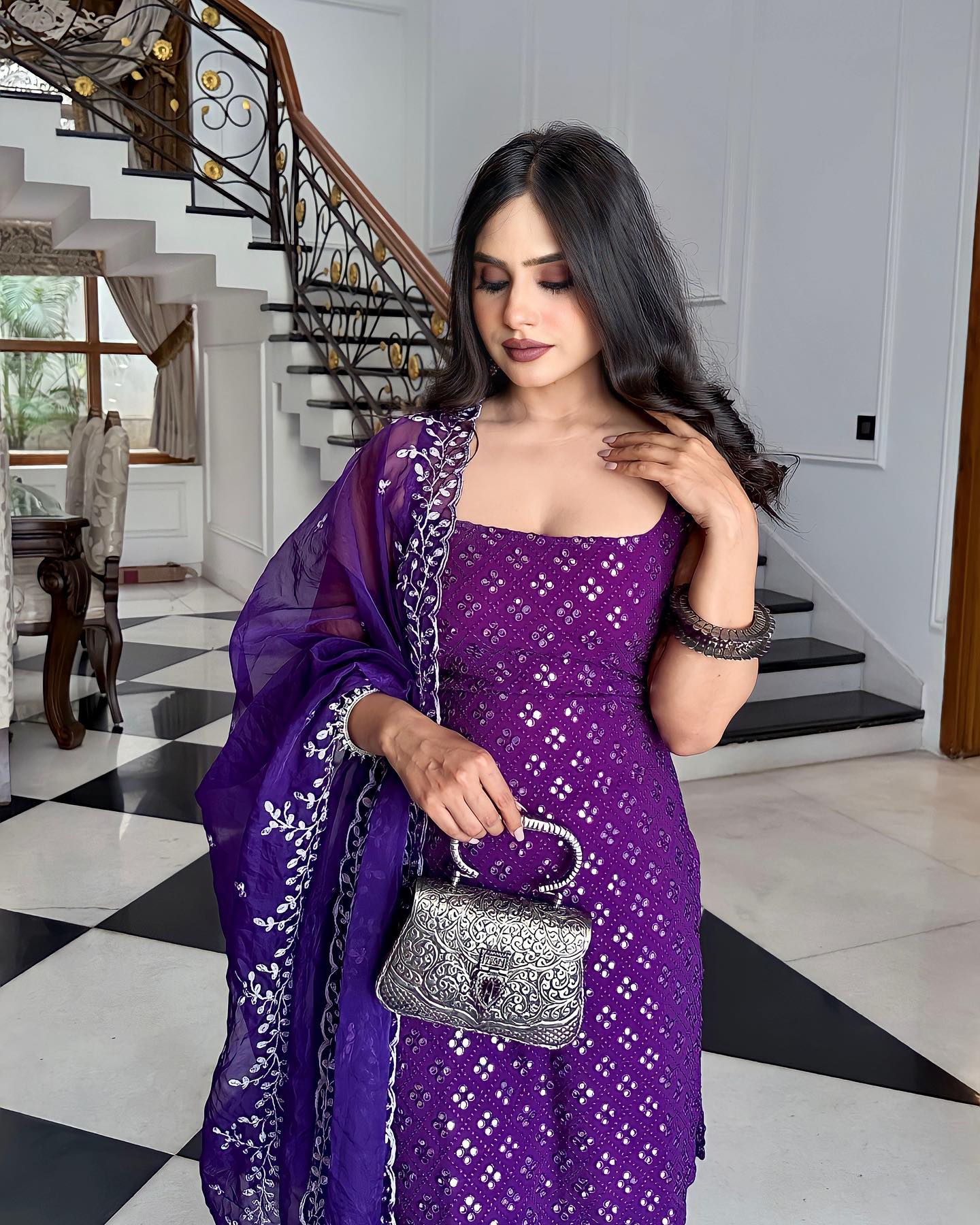 Woman in a purple saree holding a silver handbag in a home setting