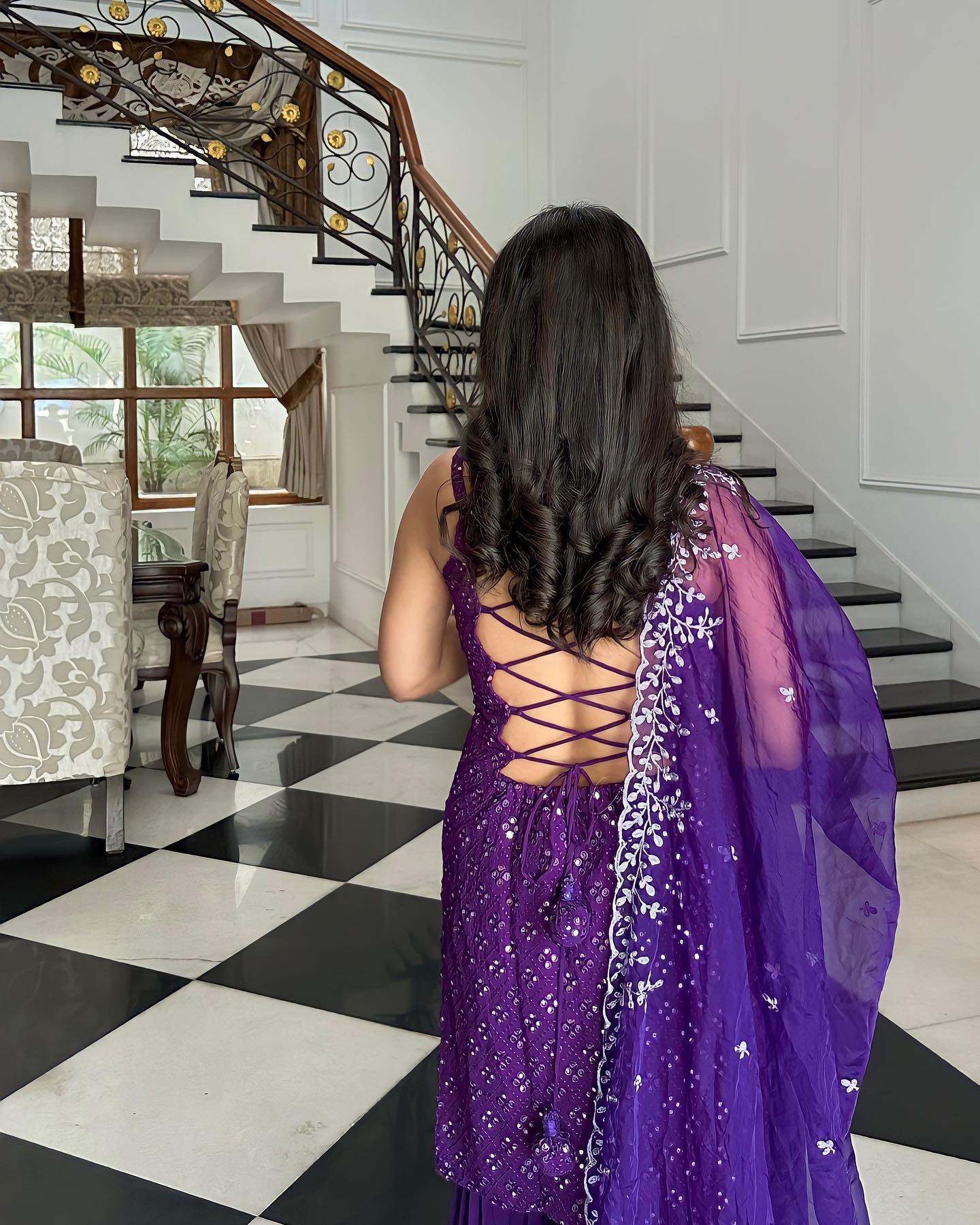 Woman in a purple saree standing in a home interior with staircase and chairs.