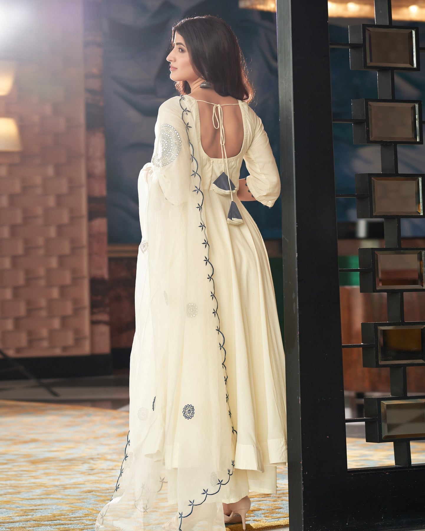 Woman in a white saree with intricate designs standing in a room with decorative walls.