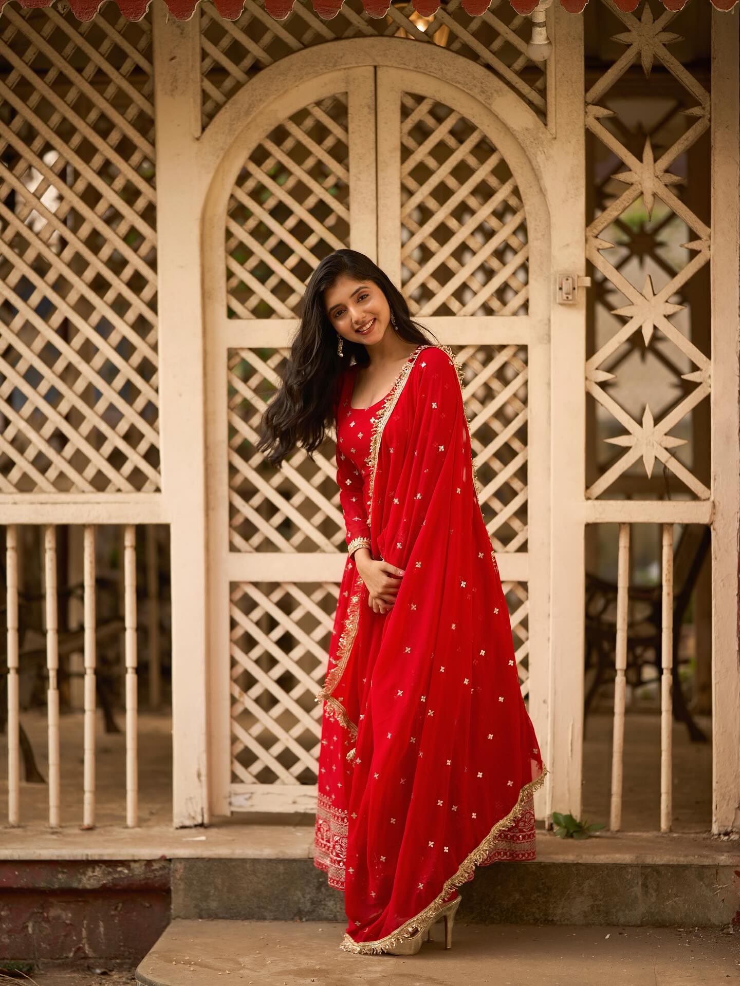 Woman in a red traditional outfit standing in front of a decorative wooden gate.