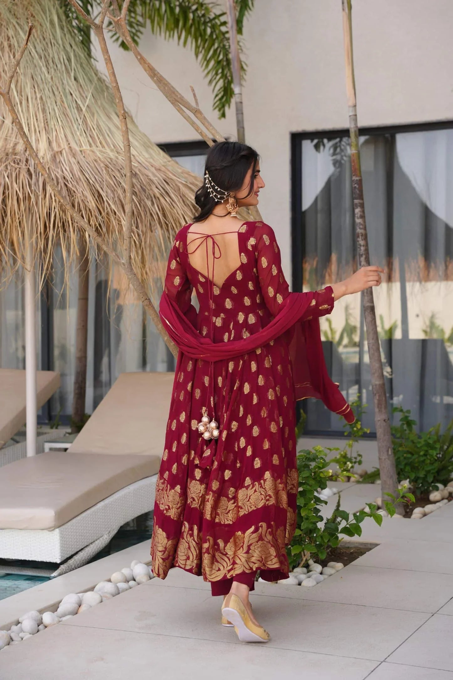 Woman in a red and gold dress standing by a poolside with palm trees and lounge chairs.