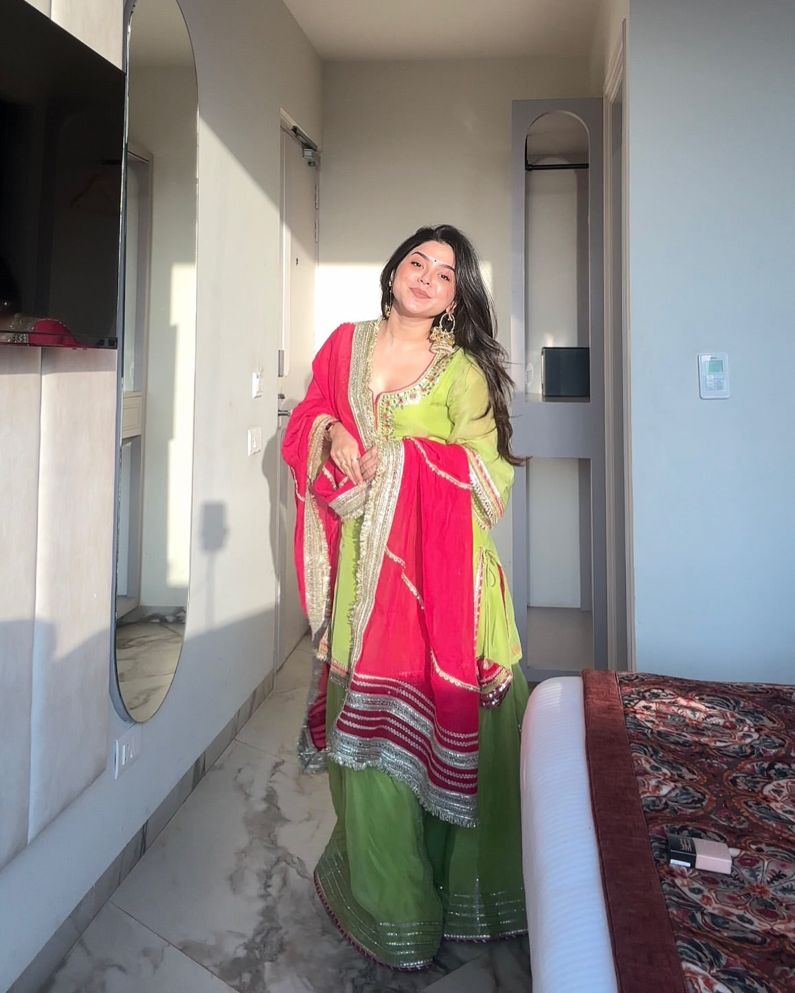 Woman in a green and pink traditional outfit standing in a room with mirrors.