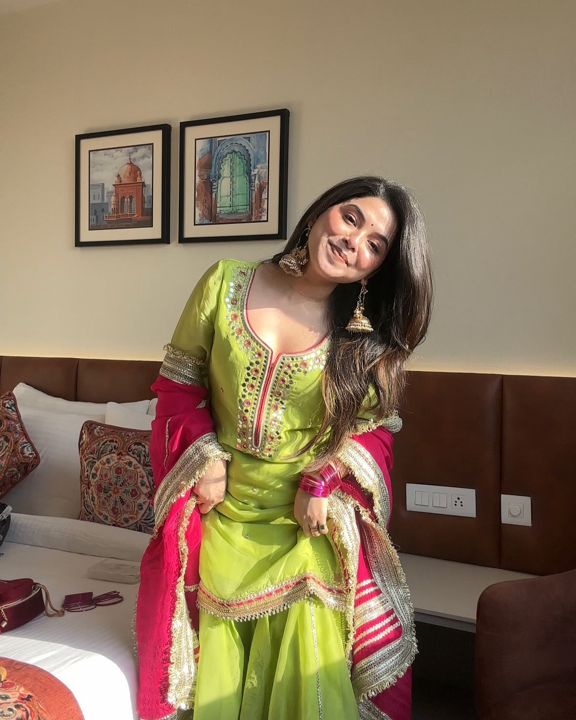 Woman in a green and pink traditional outfit posing in a room with framed artwork on the wall.