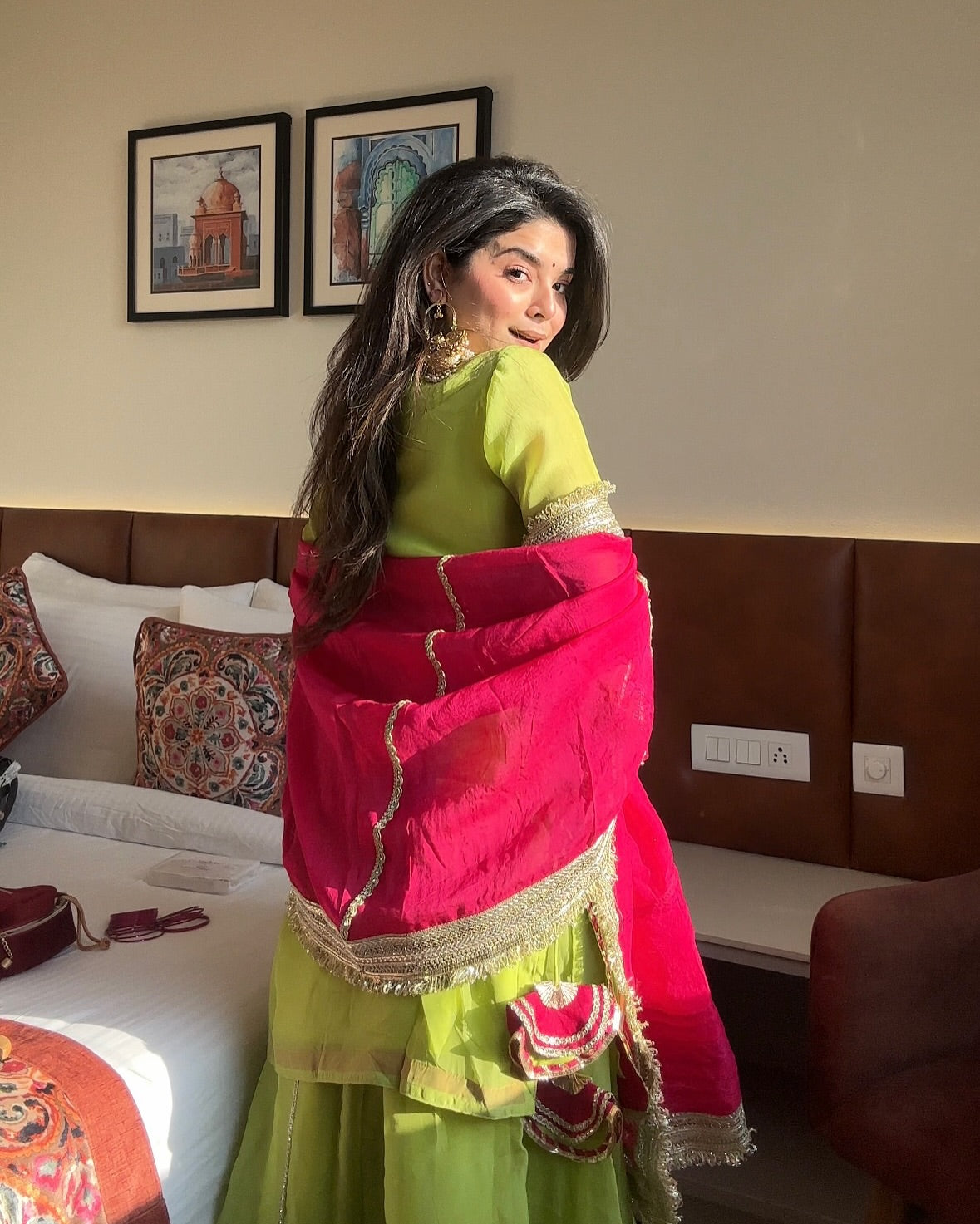 Woman in a green and pink traditional outfit standing in a room with a bed and wall art.