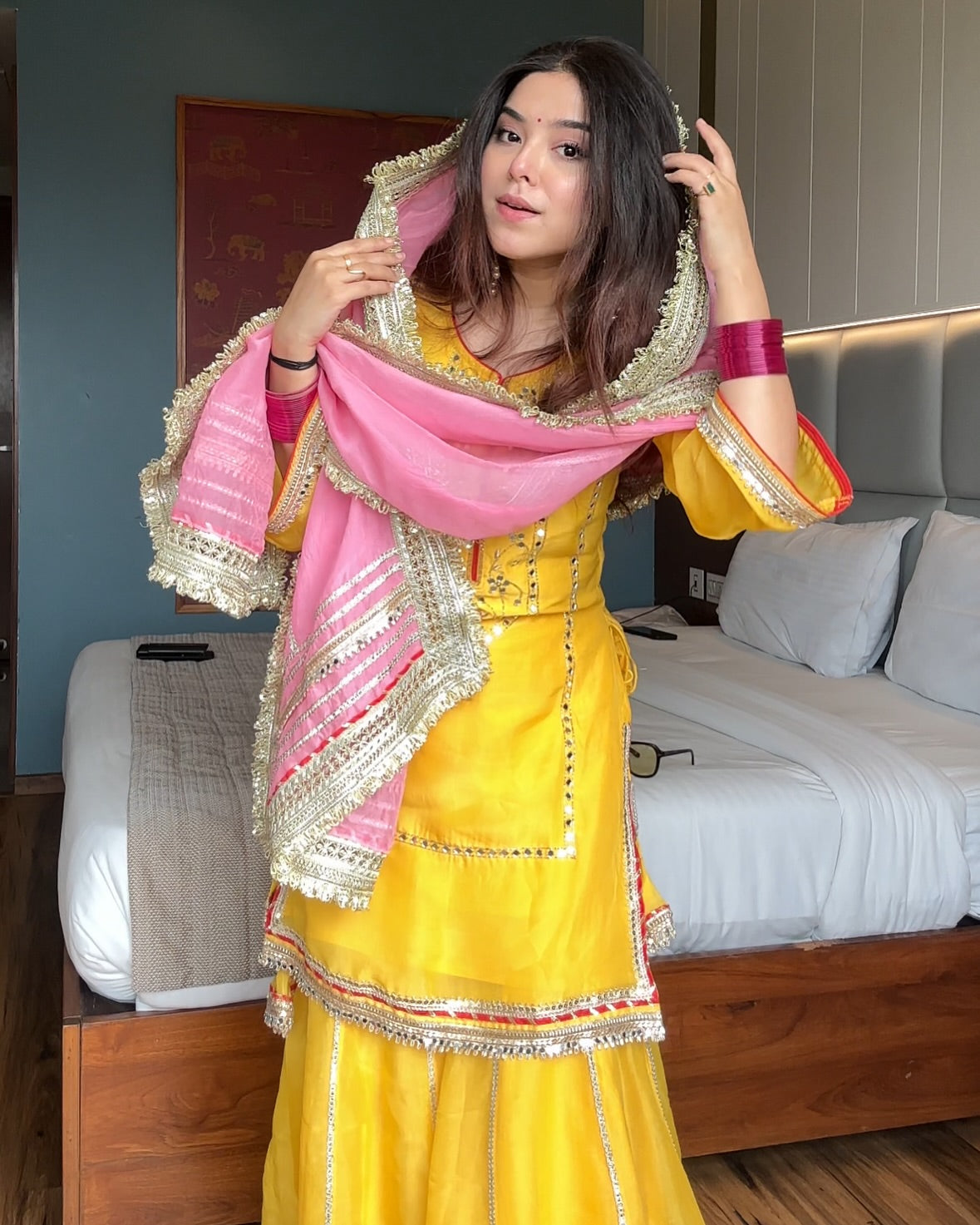 Woman in a yellow traditional outfit with a pink dupatta in a bedroom setting