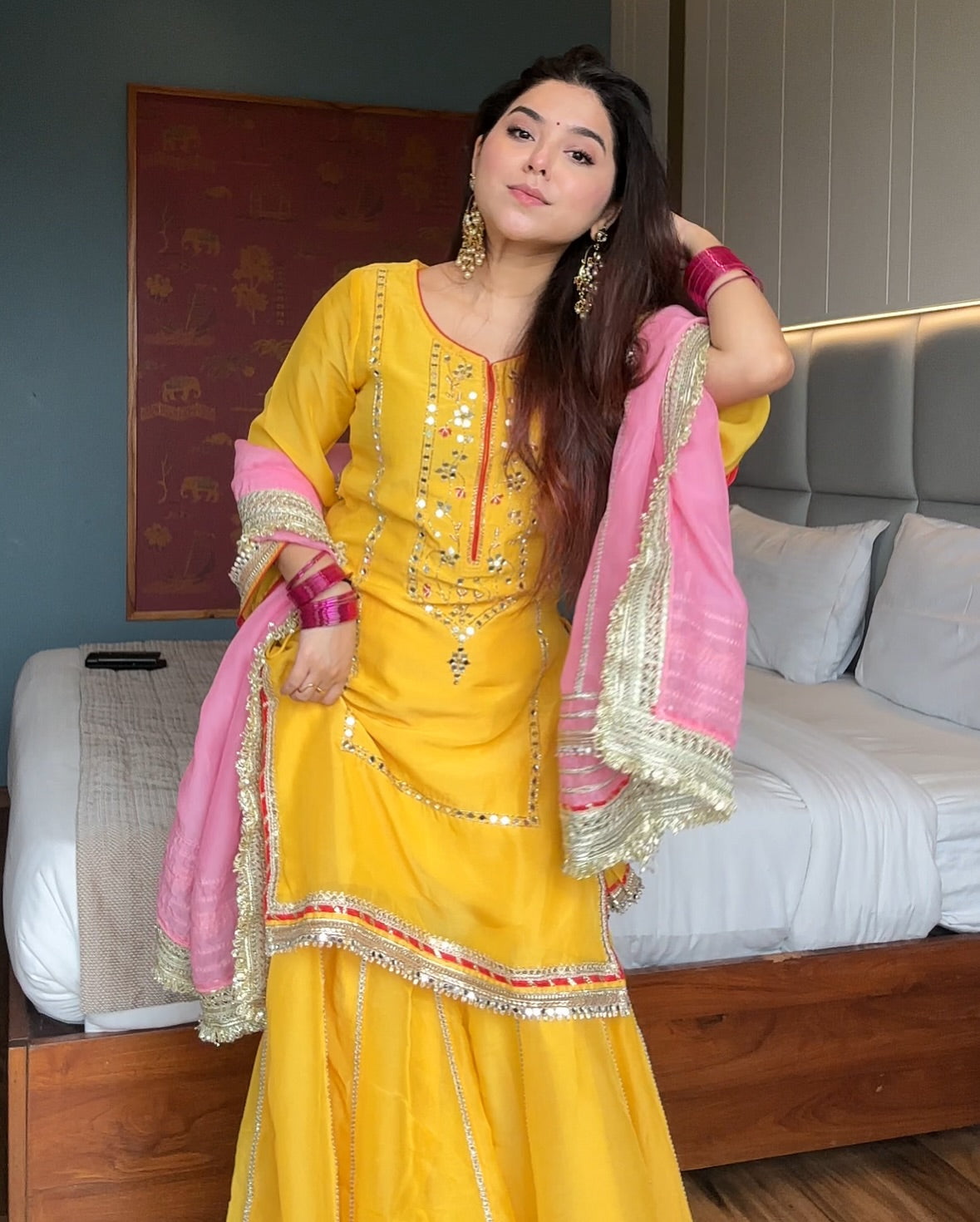 Woman in a yellow and pink traditional outfit standing in a room with a bed and wall art.