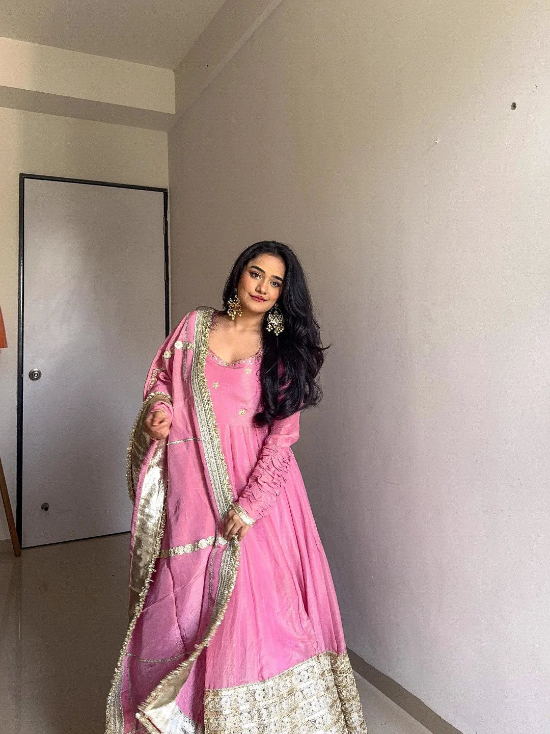 Woman in a pink traditional outfit standing indoors against a plain wall.