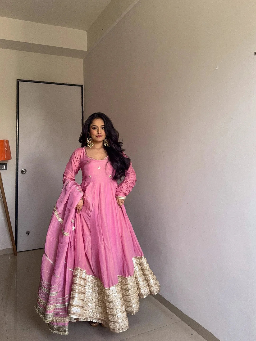 Woman in a pink traditional outfit standing in a room with a neutral wall and door.
