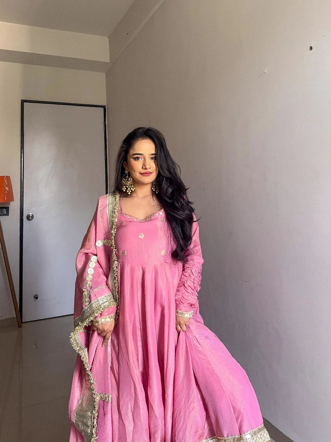 Woman in a pink traditional outfit standing indoors against a plain wall.