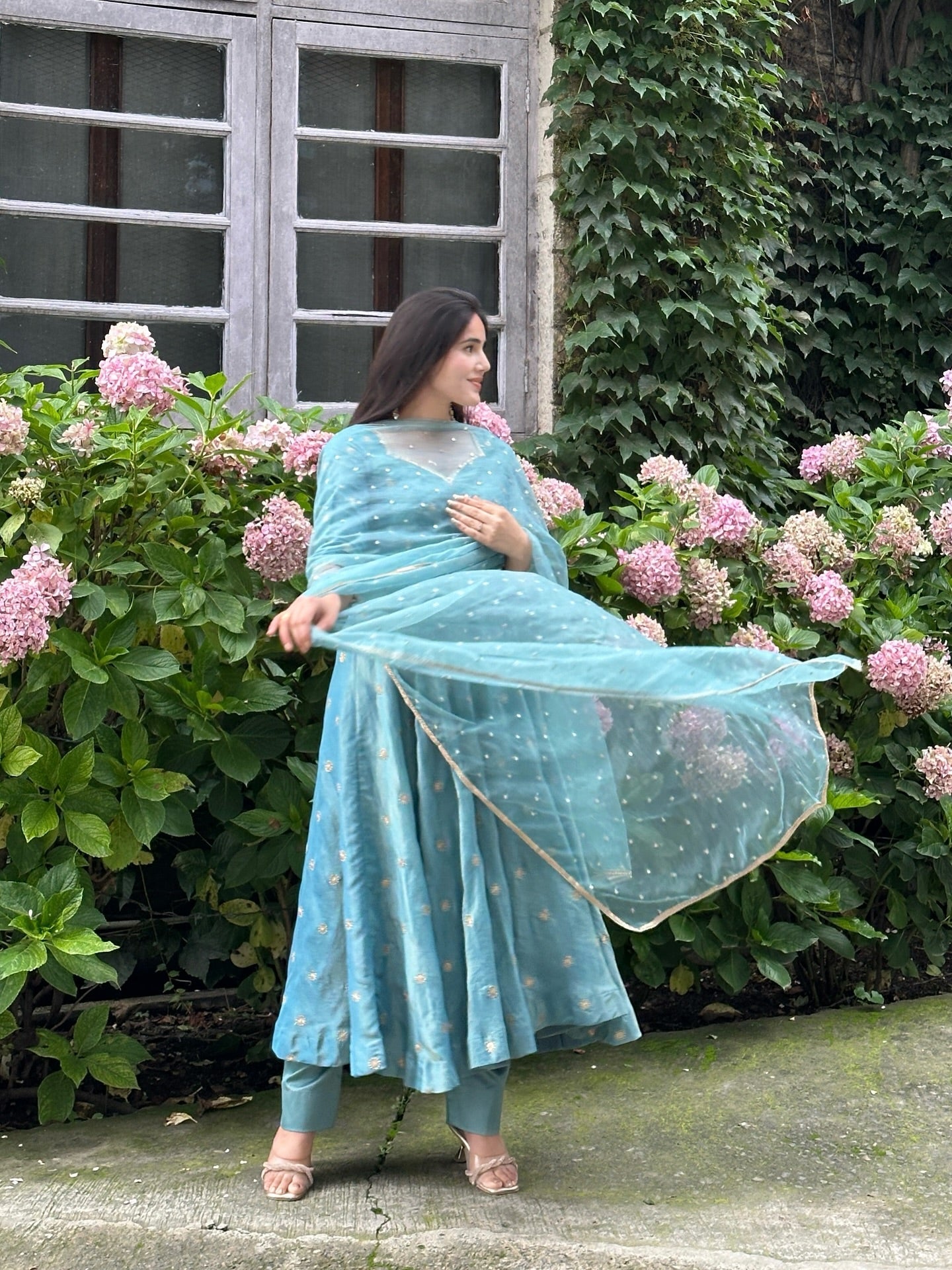 Woman in a light blue traditional outfit standing in front of pink flowers and greenery.
